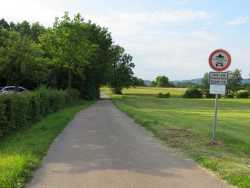 Vom Parkplatz aus führt ein asphaltierter Weg zum Starkholzbacher See und um den See. Rechts davon sind Felder. Man sieht auch die offene Schranke.